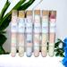 Whole Self Aromatherapy's bath salts in test tubes on a white towel, with greenery in the background and a blue flower to the right.